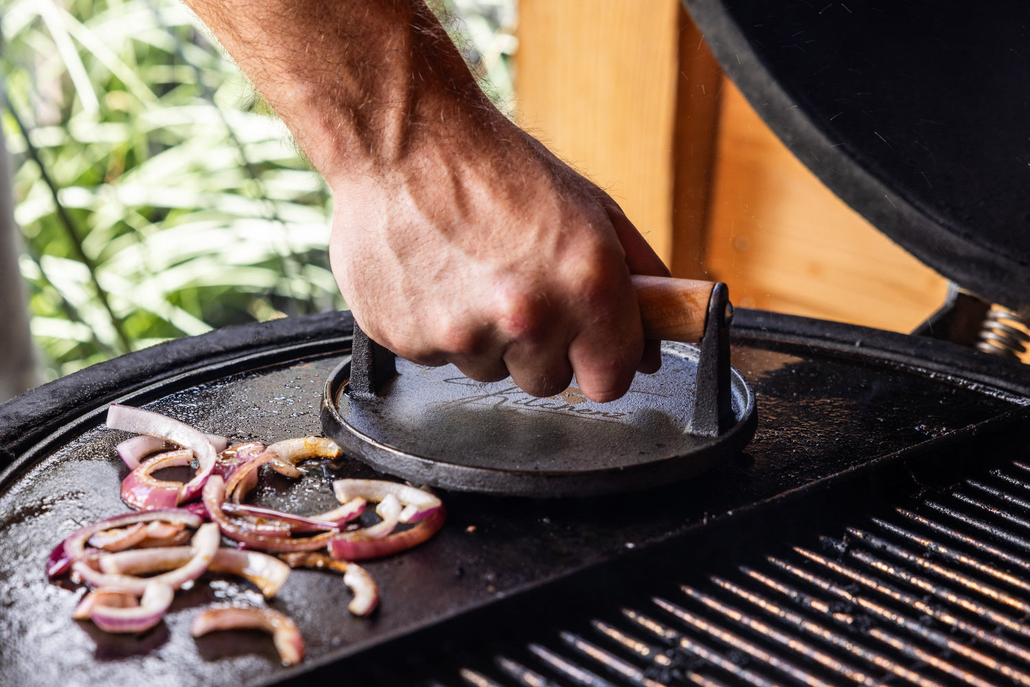 Ronde gietijzeren hamburgerpers van Kitchenz met houten handvat wordt gebruikt op een kamado barbecue grill om uien en burgers te grillen. ronde hamburgerpers, gietijzeren vleespers, smash burger tool, Kitchenz BBQ accessoires, kamado grill gereedschap, vleesaandrukker gietijzer.