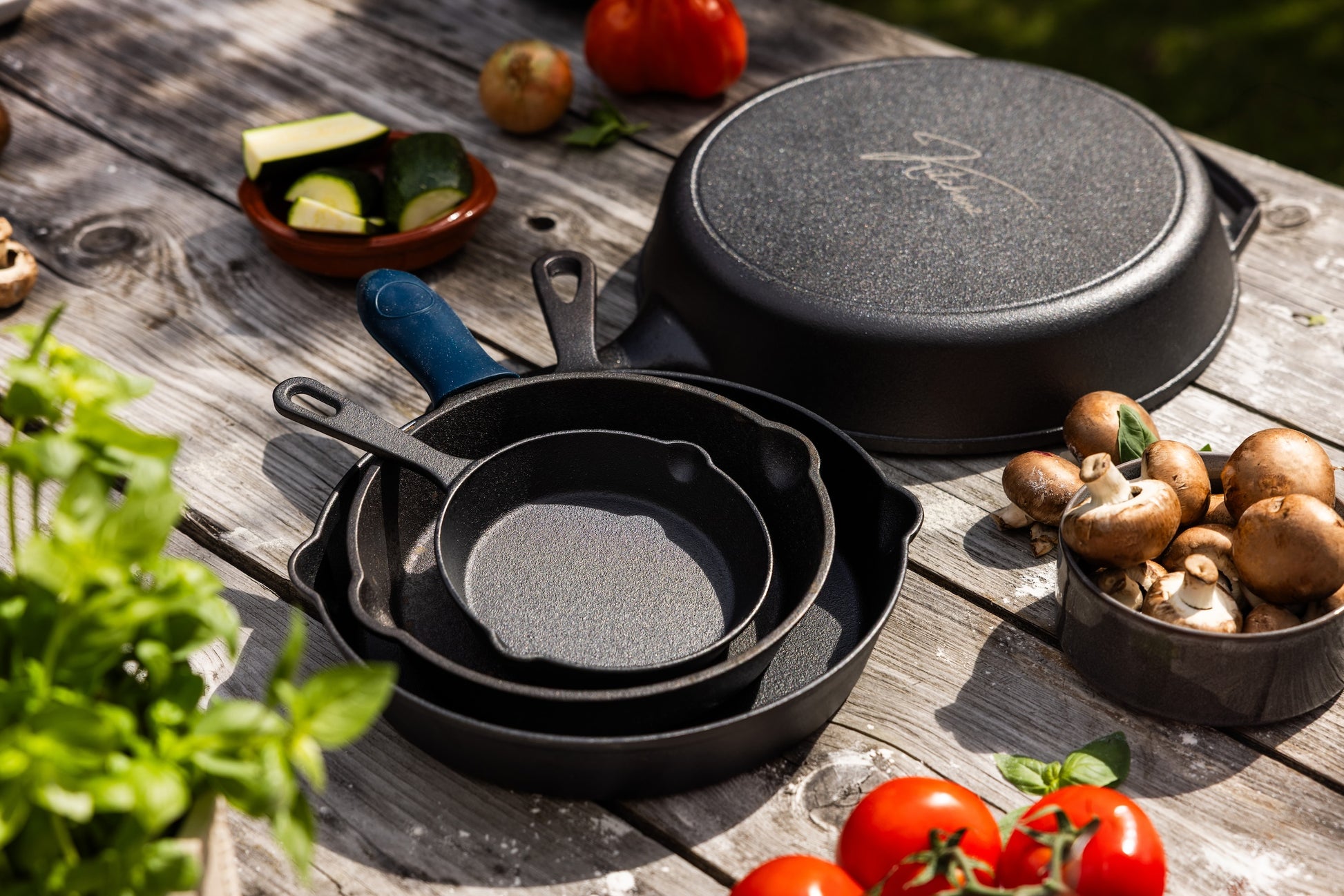 Een complete set van zwarte gietijzeren pannen van Kitchenz in de maten 15cm, 20cm, 25cm en de grote 30cm XL skillet op een houten tafel met verse groenten.  gietijzeren skillet set, Kitchenz pannenset gietijzer, koekenpan inductie set, kleine en grote skillet, gietijzeren pan 30cm, bbq pannenset.