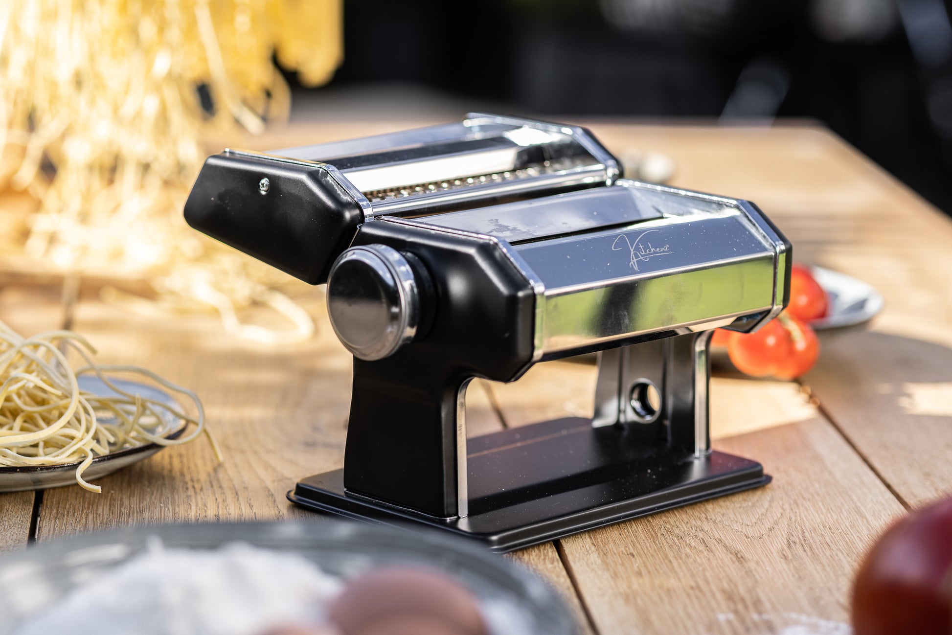 Een close-up van een stijlvolle, zwartgecoate pastamachine met een minimalistisch industrieel ontwerp, voorzien van een contrasterende metalen instelknop voor de deegdikte. 
zwarte pastawals, Kitchenz matte pastamachine, modern keukendesign zwart, luxe deegroller machine, donkere uitvoering pastamaker, Kitchenz noir kookgerei.