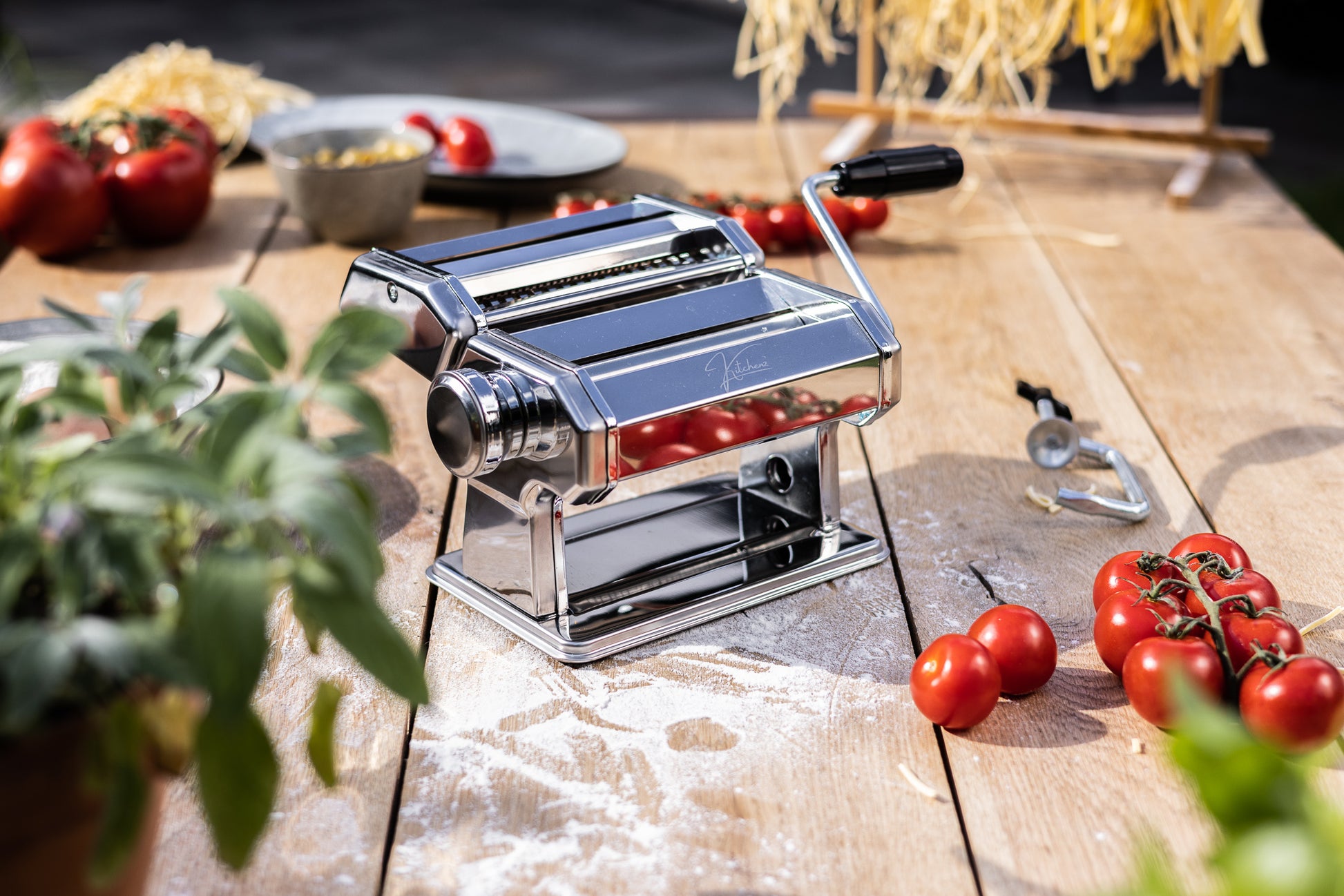 Een glanzende, zilverkleurige pastamachine van Kitchenz met een spiegelende finish en een houten zwengel, gepositioneerd op een marmeren keukenblad. 
chromen pastamaker, Kitchenz zilveren pasta wals, roestvrijstalen deegroller, authentieke Italiaanse pastamachine, glimmend keukenapparaat, handmatige tagliatelle snijder.