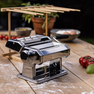 Een complete set bestaande uit een glanzende zilveren pastamachine naast een houten droogrek met acht armen waaraan verse slierten tagliatelle hangen te drogen op een aanrecht. 
pastamachine met droogrek, Kitchenz pastaset chroom, houten deegdroger set, verse pasta maken pakket, Kitchenz keukenhulp bundel, complete pastasnijder set.