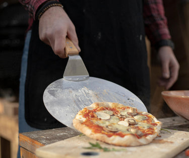Een scherpe studio-opname van een roestvrijstalen Kitchenz pizzaschep met een houten handvat, liggend op een marmeren werkblad naast wat vers gestrooid meel.
professionele pizzaschep, Kitchenz rvs bakspatel, pizzaschuiver met houten grip, keukenhulp voor steenoven, Kitchenz authentieke pizzaschep, metalen deegschep.