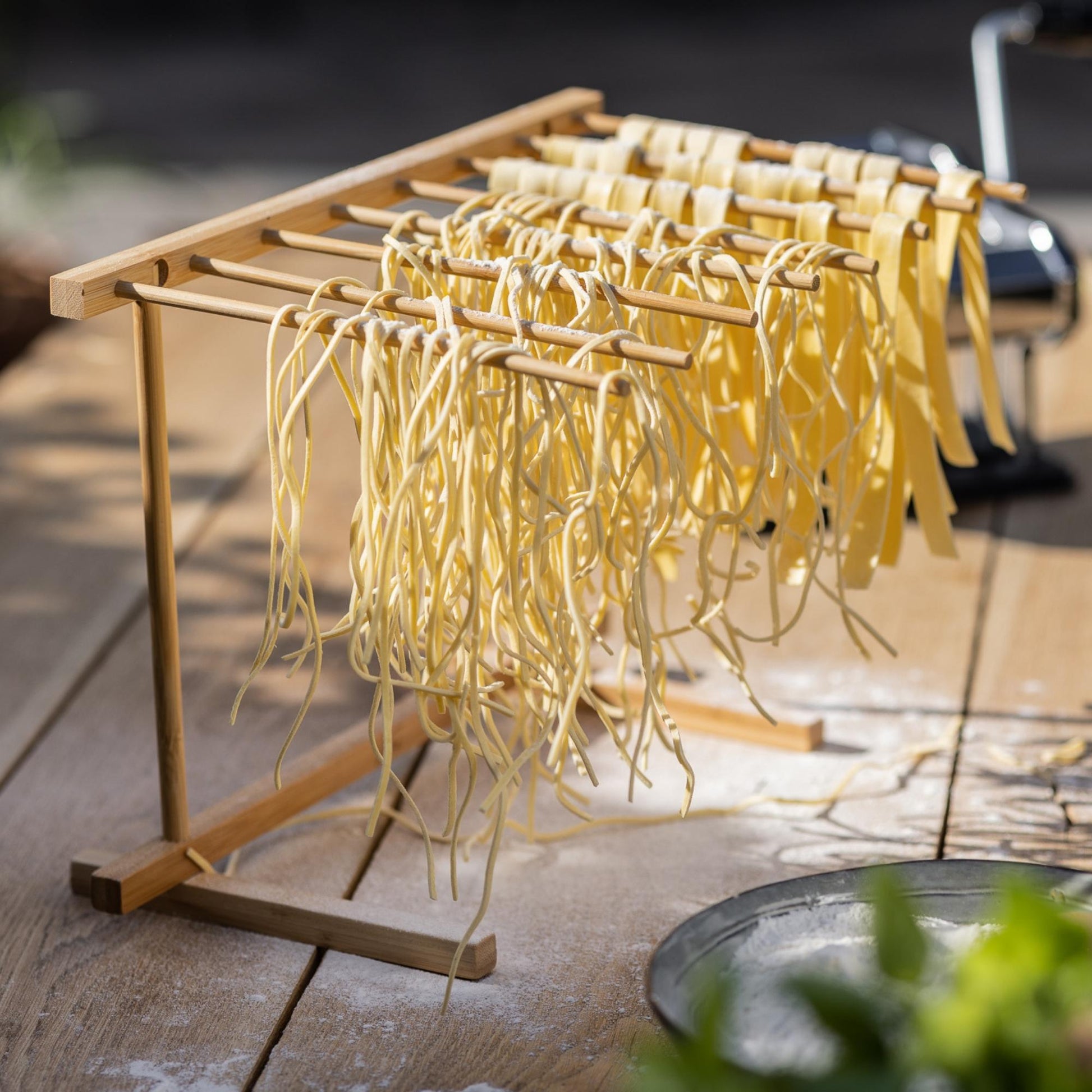 Een bovenaanzicht van een glimmende Kitchenz pastamachine in actie, waarbij verse deegslierten direct vanuit de machine op de houten sporten van het compacte droogrek worden gelegd.
Kitchenz pastamachine set, verticaal pastadroogrek, complete deegroller bundel, verse spaghetti maken, Kitchenz keukenaccessoires, handmatige pastawals met standaard.